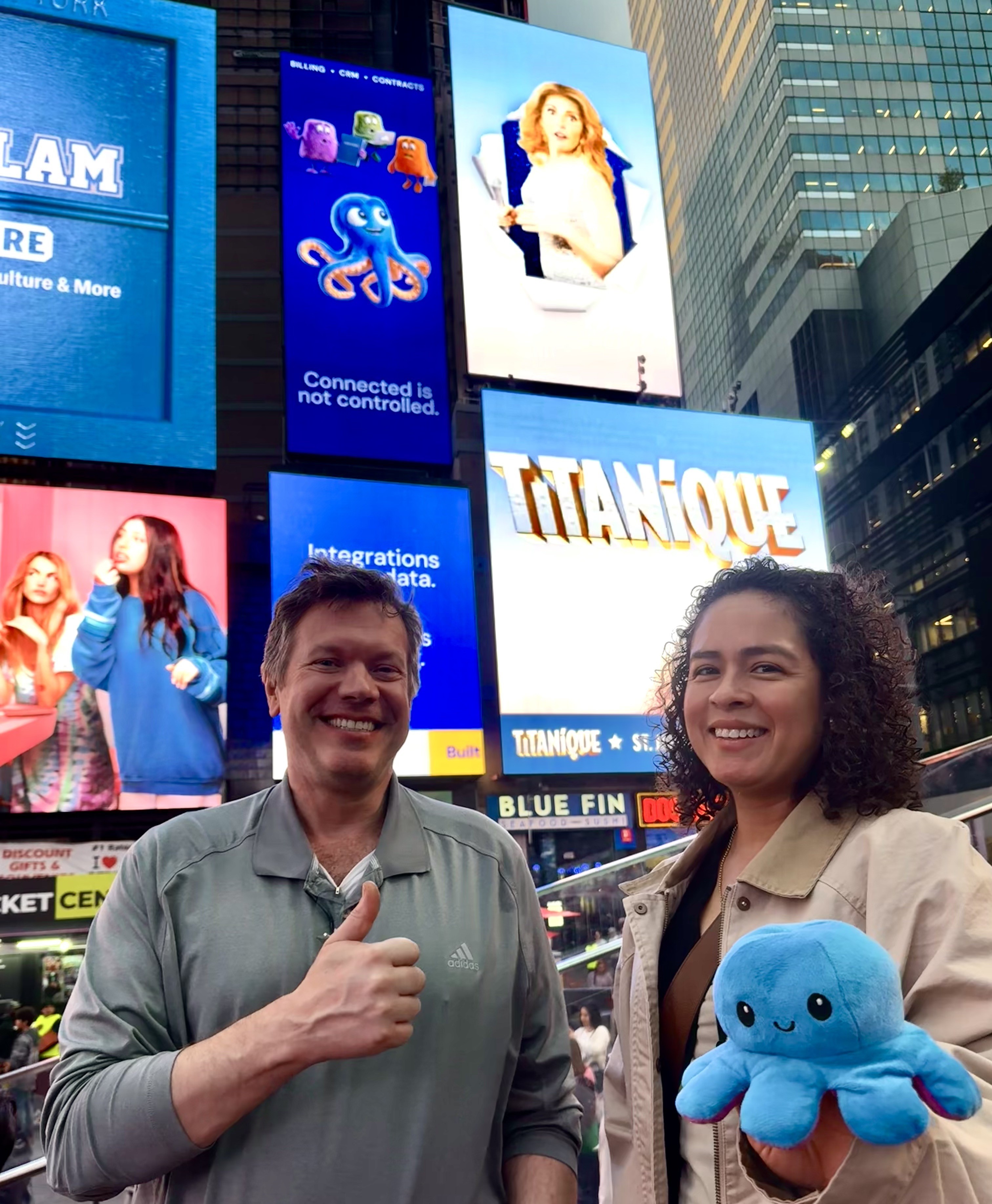 Sandra and JW in Times Square, standing under the Meshly billboard, Sandra holding a plush octopus